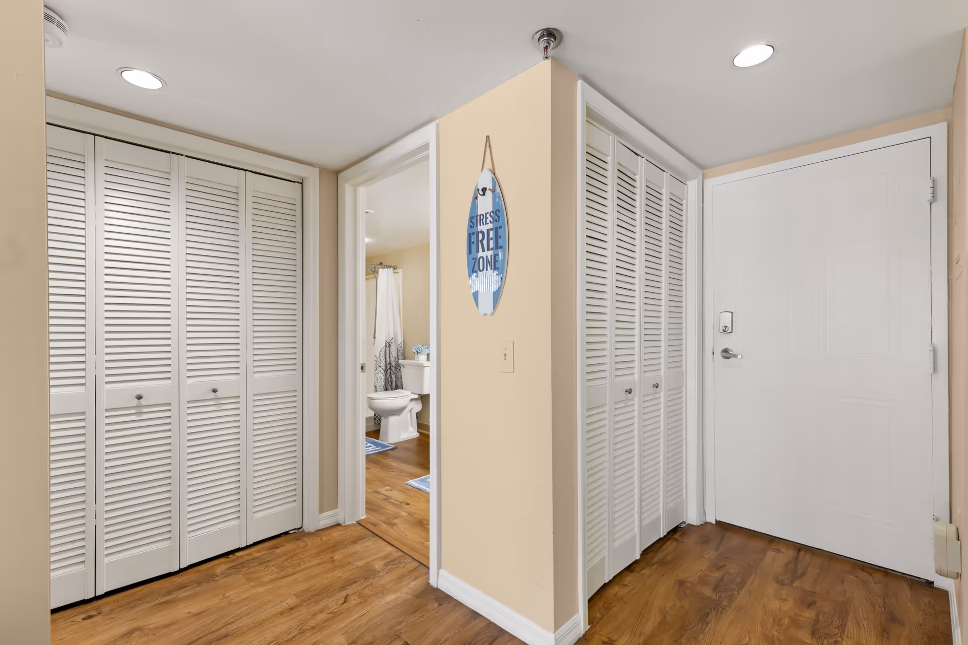 Interior view of a hallway in a senior living facility with light beige walls and wood flooring. There are two sets of white louvered closet doors on either side of the hallway. A white entrance door with a keypad lock is visible on the right. Through an open doorway, a bathroom with a toilet, shower curtain, and blue bath mats can be seen. A decorative sign hanging on the wall reads 'Stress Free Zone'.