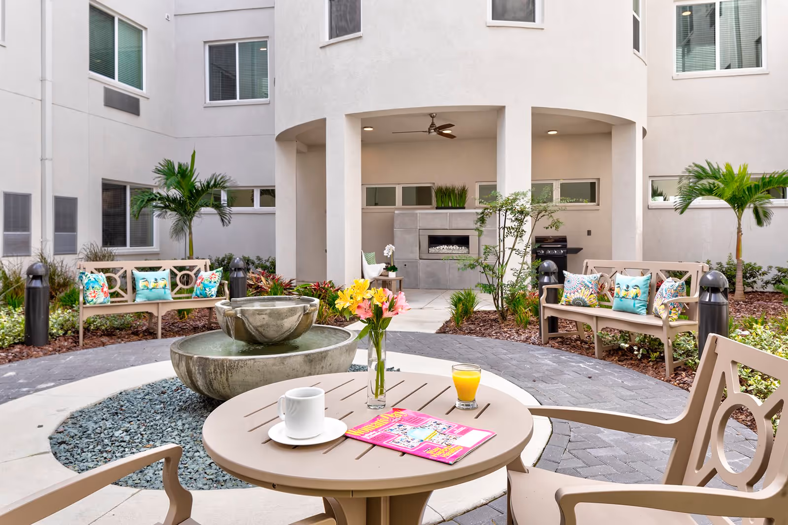 Outdoor courtyard area with a round table and chairs in the foreground, featuring a cup of coffee, a glass of orange juice, a vase with flowers, and a magazine on the table. In the background, there are benches with colorful cushions, palm plants, a water fountain, and a covered patio area with a ceiling fan and a fireplace.