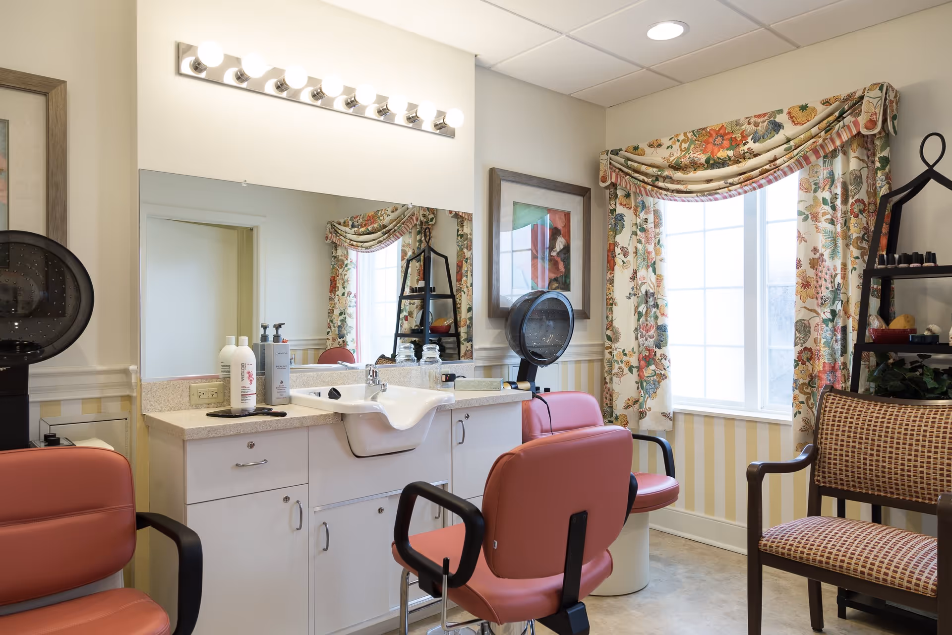 A bright salon room with two pink salon chairs, a white sink, a large mirror with lights above it, floral curtains on the window, a framed picture on the wall, and a black shelving unit holding various hair and nail products.
