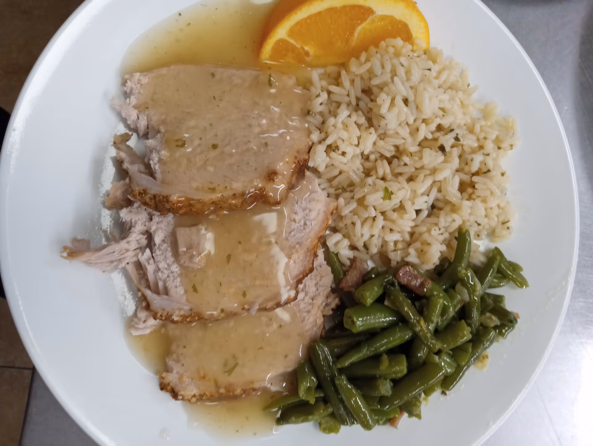 A white plate with three slices of turkey covered in gravy, a serving of cooked rice, green beans with bits of bacon, and a slice of orange as garnish.