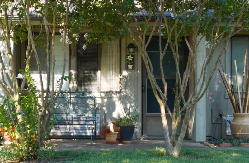 Front porch of a retirement community unit with a bench, potted plants, trees, and a screen door.