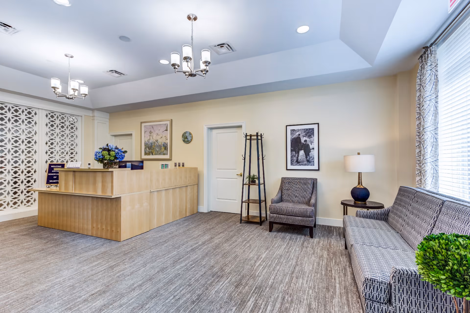 Reception area of Artis Senior Living of Wilmette featuring a wooden front desk with a flower arrangement, a welcome sign, two armchairs, a sofa, a side table with a lamp, framed artwork on the walls, and large windows with patterned curtains allowing natural light.