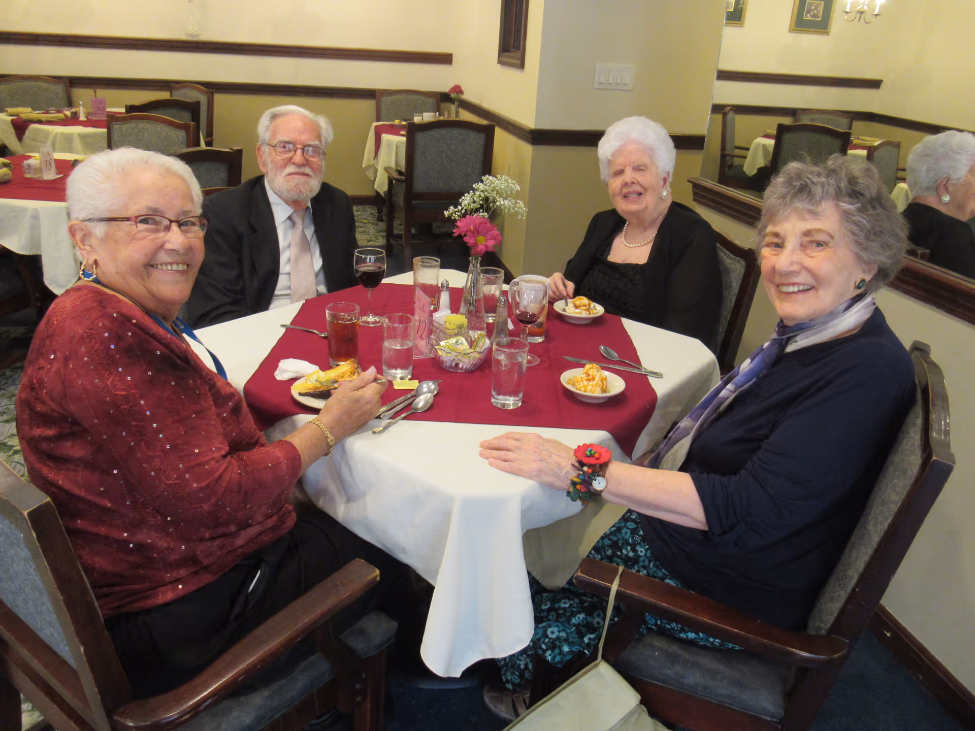 Four elderly people sitting around a round dining table in a dining room, smiling and enjoying dessert. The table is covered with a white tablecloth and a red table runner, with glasses of water and wine, silverware, and a small flower arrangement in the center. The background shows other tables with similar settings and chairs.