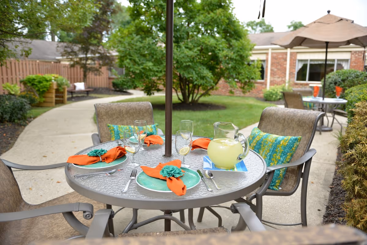 Outdoor patio area with a round glass table set for four, featuring green plates, orange napkins with teal napkin rings, and glasses with lemon slices. A pitcher of lemonade is on the table. The table is surrounded by cushioned chairs with colorful pillows, and there are umbrellas providing shade. In the background, there is a brick building, green bushes, trees, and a paved walkway.