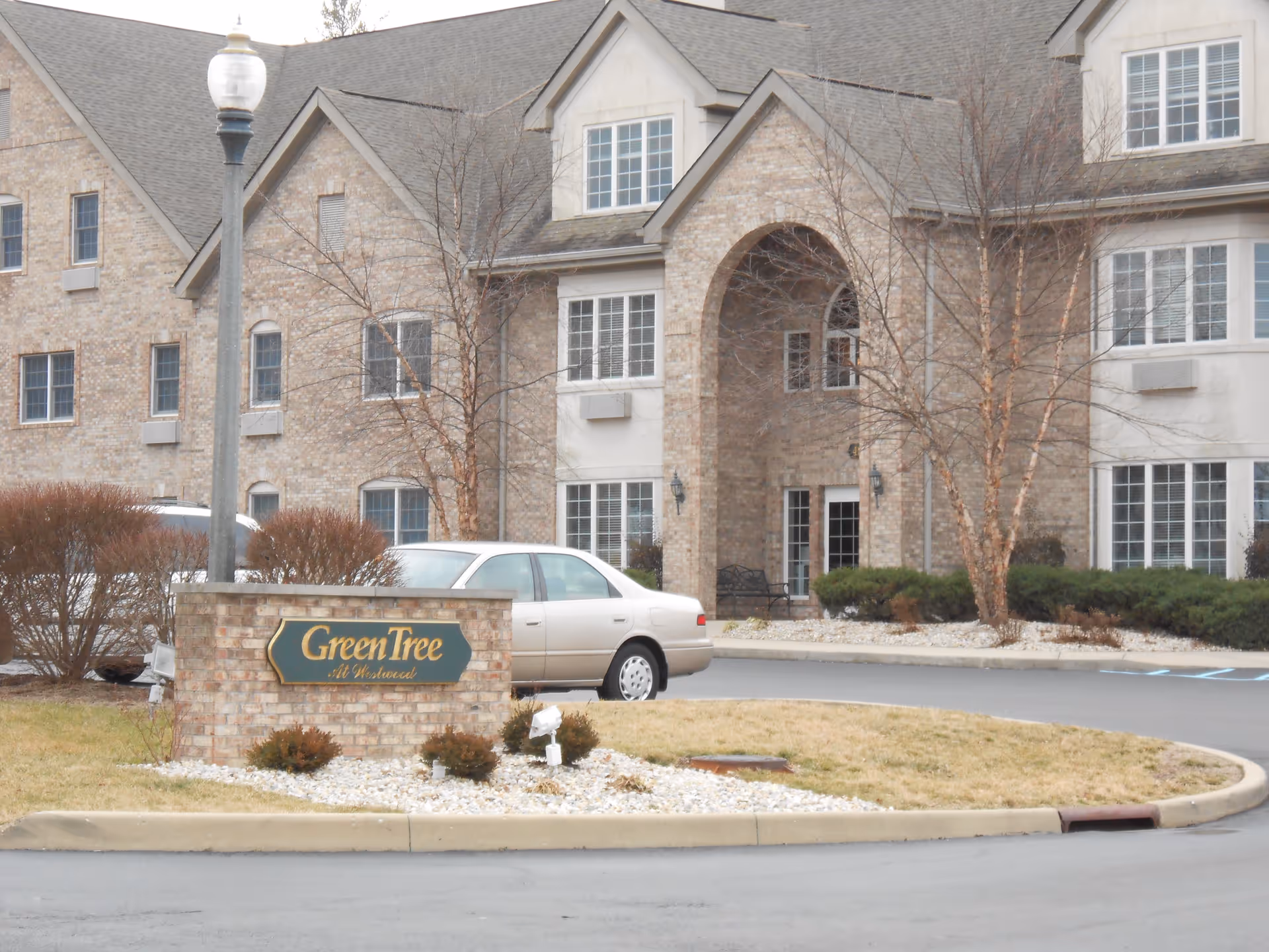 Exterior view of a senior living facility named GreenTree At Westwood, showing a brick building with multiple windows, an arched entrance, a parked beige car, and a sign with the facility name on a brick base surrounded by landscaping.