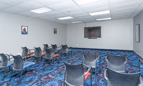 A small conference or meeting room with rows of black chairs facing a blank wall with a mounted TV screen. The room has a patterned blue and purple carpet, white walls with a few framed pictures, and a ceiling with fluorescent lights.