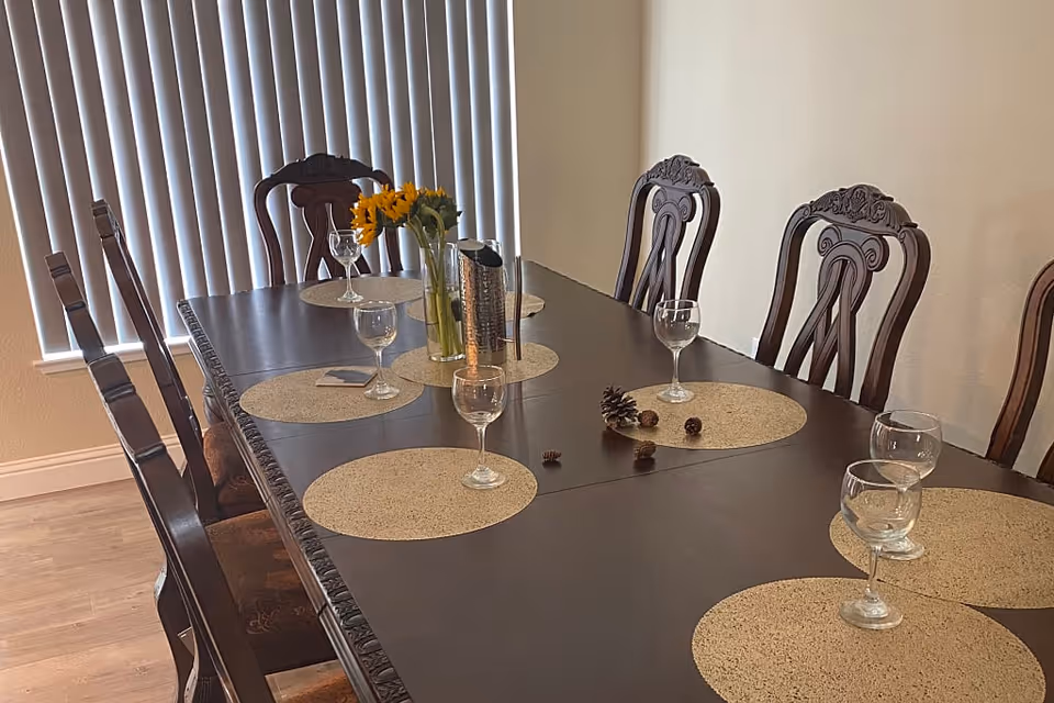 Long dark wood dining table with round placemats, wine glasses, a vase of sunflowers and wooden chairs in a room with vertical blinds.