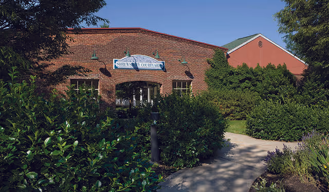 Exterior view of a brick building with a sign that reads 'Shrewsbury Courtyards' above the entrance. The building is surrounded by lush green bushes and trees, with a paved walkway leading to the entrance under a clear blue sky.
