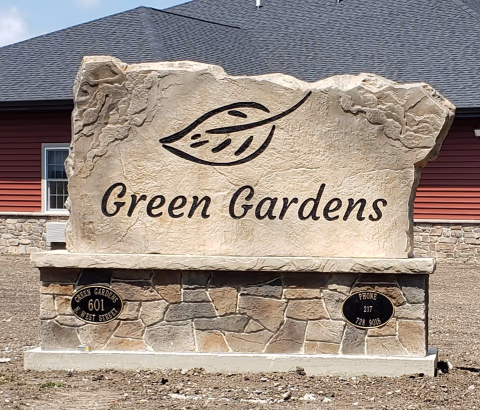 A large stone sign with a carved leaf design and the words 'Green Gardens' engraved on it, set in front of a building with red siding and stone accents.