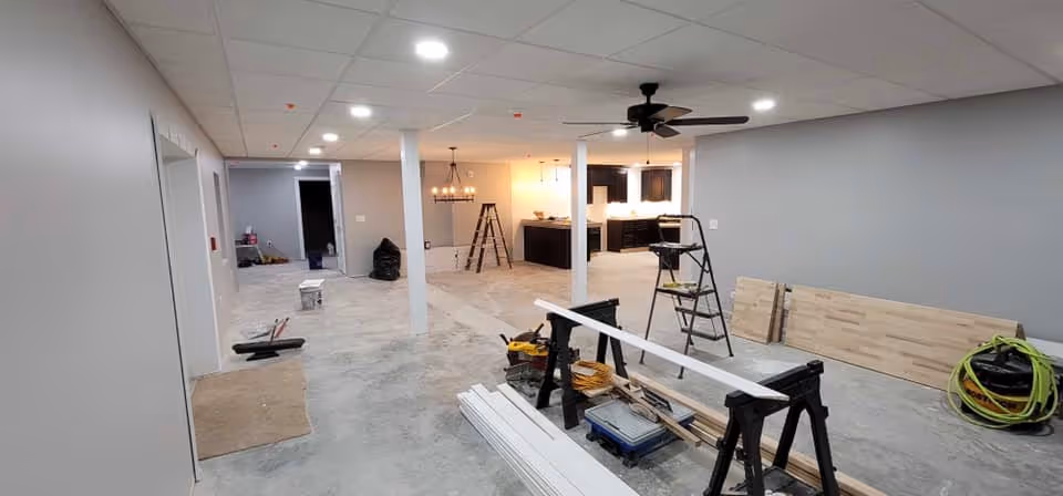 Interior view of a large room under renovation with construction materials and tools scattered around. The room has gray walls, a ceiling fan, recessed lighting, and an open kitchen area with dark cabinets and pendant lights in the background. There are ladders, wooden planks, and various equipment on the concrete floor.