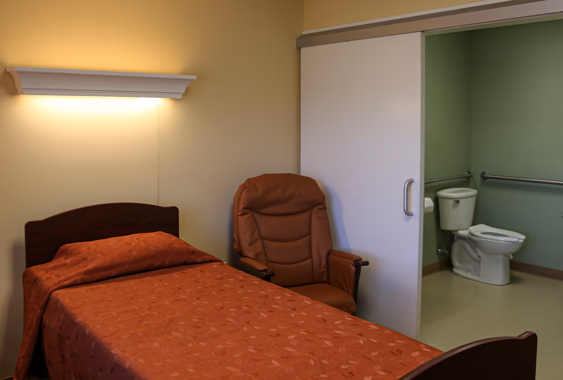 A small resident bedroom with a single bed, a recliner chair, and an open accessible bathroom showing a toilet.
