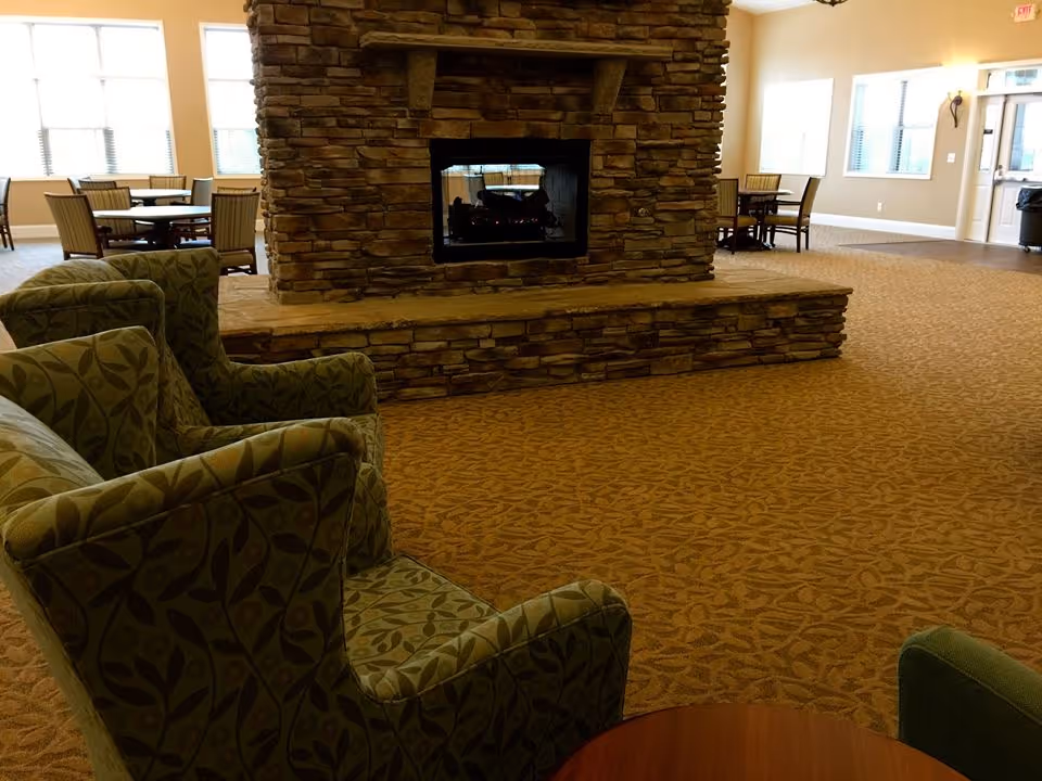 A spacious common area with a large stone fireplace in the center. There are patterned armchairs in the foreground and round tables with chairs near large windows in the background. The room has beige walls, carpeted floors with a leaf pattern, and ample natural light.