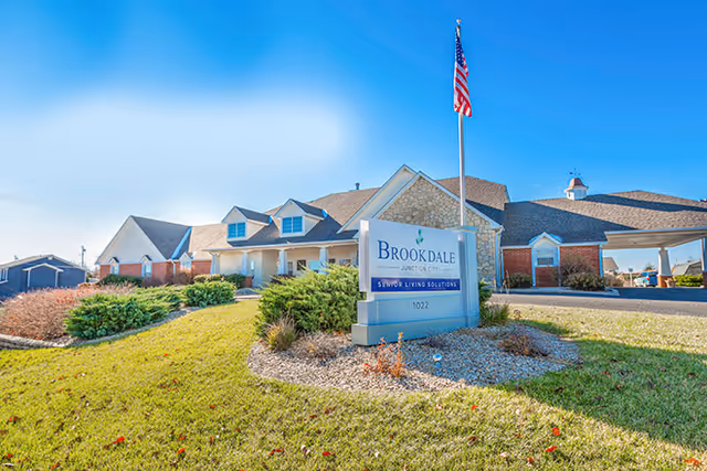 Front exterior of the Brookdale Junction City senior living building with a sign, American flag, and landscaped lawn.