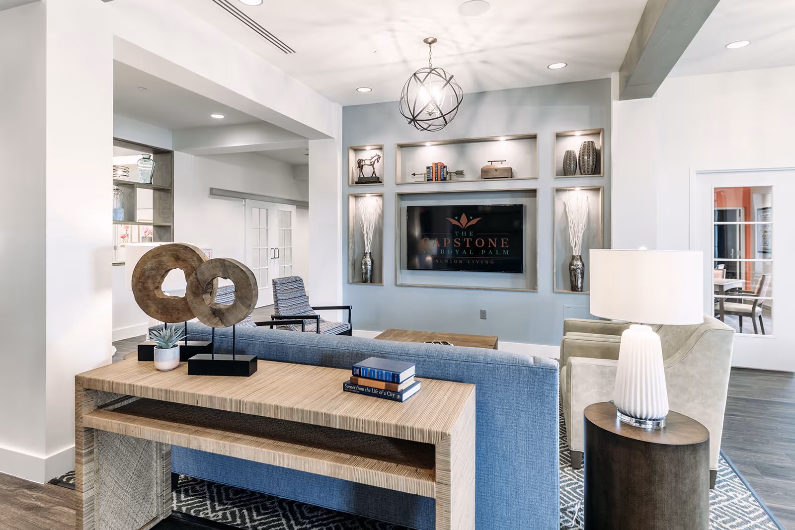 Bright, modern lounge with a sofa, armchairs, a console table, and a built-in TV wall with shelving and decorative accents.