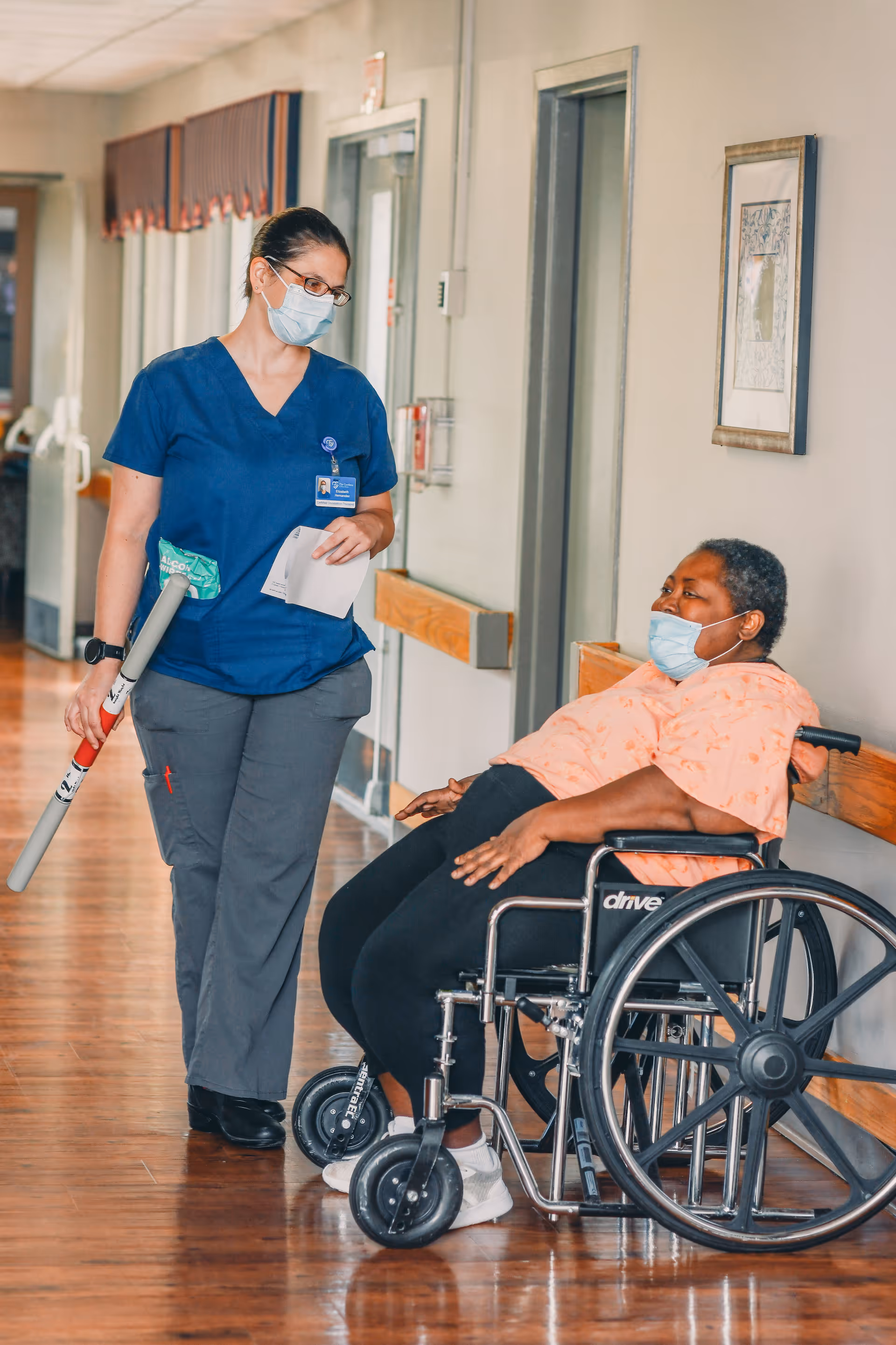 A healthcare worker wearing blue scrubs and a face mask is standing and talking to a woman in a wheelchair who is also wearing a face mask. They are in a hallway with wooden floors and light-colored walls, with a framed picture on the wall and doors in the background.