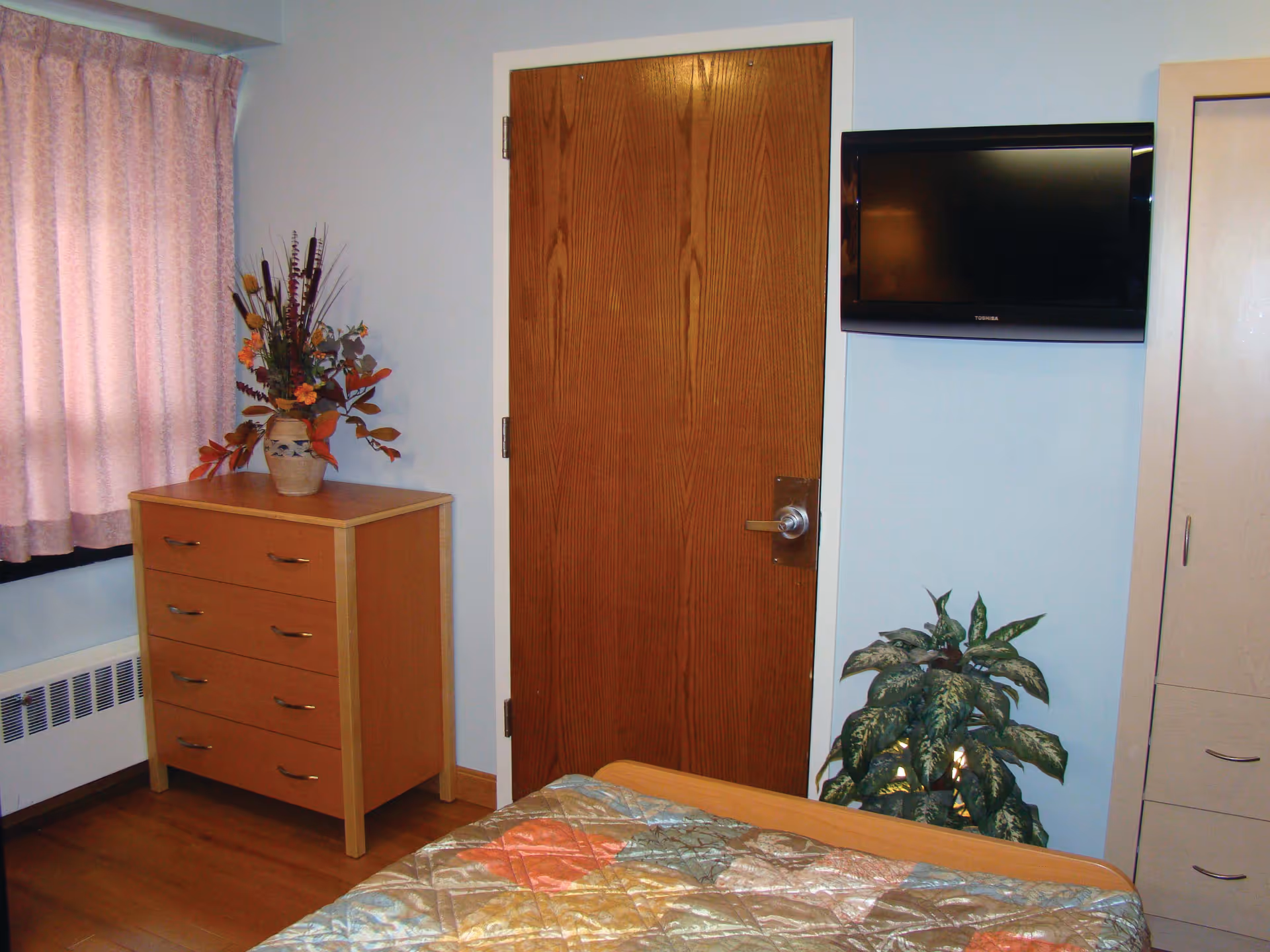A small bedroom with a wooden door, a chest of drawers with a vase of flowers on top, a mounted flat-screen TV, a leafy potted plant, and part of a bed with a colorful quilted bedspread. Pink curtains cover a window on the left side.