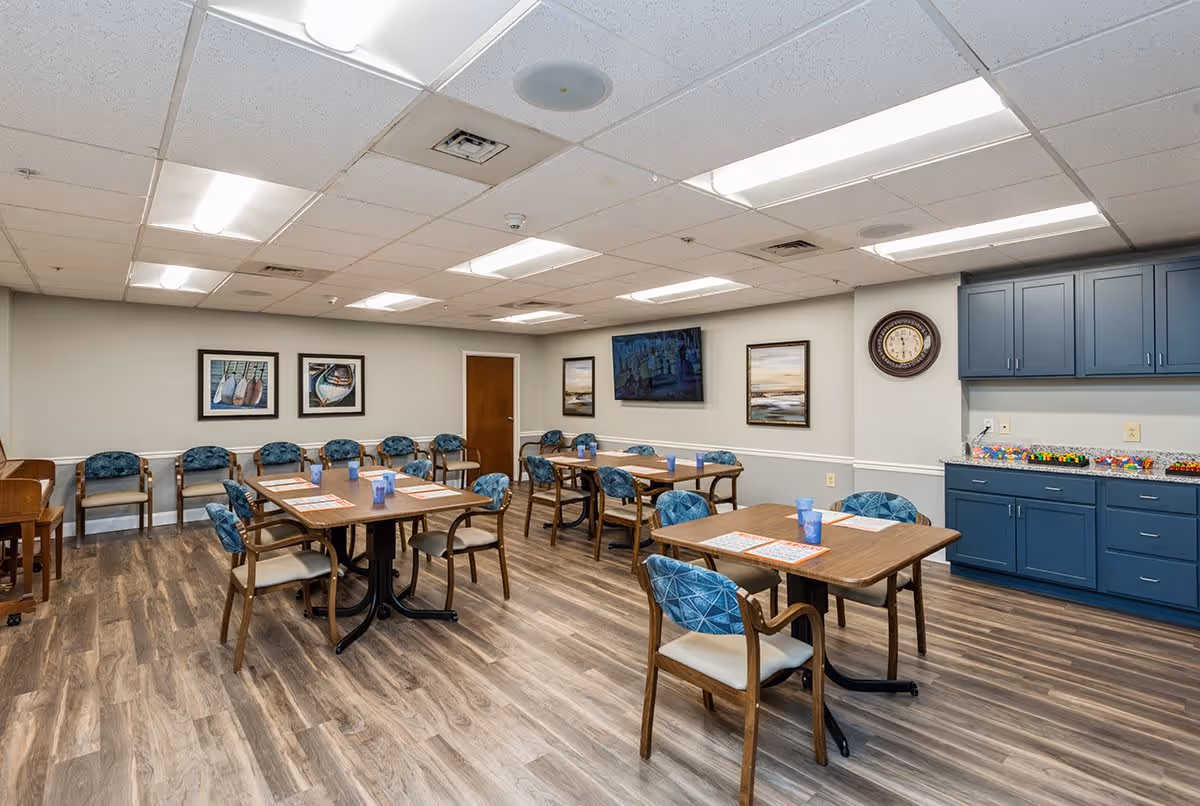 A well-lit dining room with several wooden tables and blue cushioned chairs arranged neatly. The room has wood-patterned flooring, blue cabinets on the right side with various items on the countertop, a wall clock, framed artwork, and a flat-screen TV mounted on the wall. Additional chairs are lined up against the far wall.