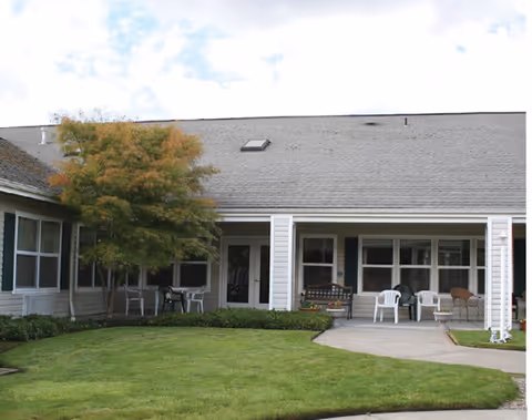 Front exterior of a one-story senior living building with a covered porch, lawn, and patio chairs.