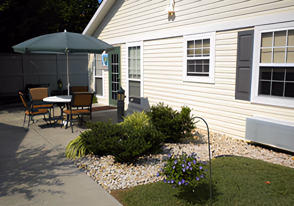 Outdoor patio area next to a light yellow building with white trim and windows. The patio has a round table with four chairs and a large green umbrella providing shade. There are bushes and a small flower bed with purple flowers near the building, and a concrete walkway leading to a green door.