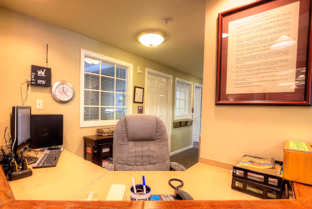 Reception desk and office chair in an interior lobby area with a computer monitor, framed document, wall clock, and a hallway beyond.