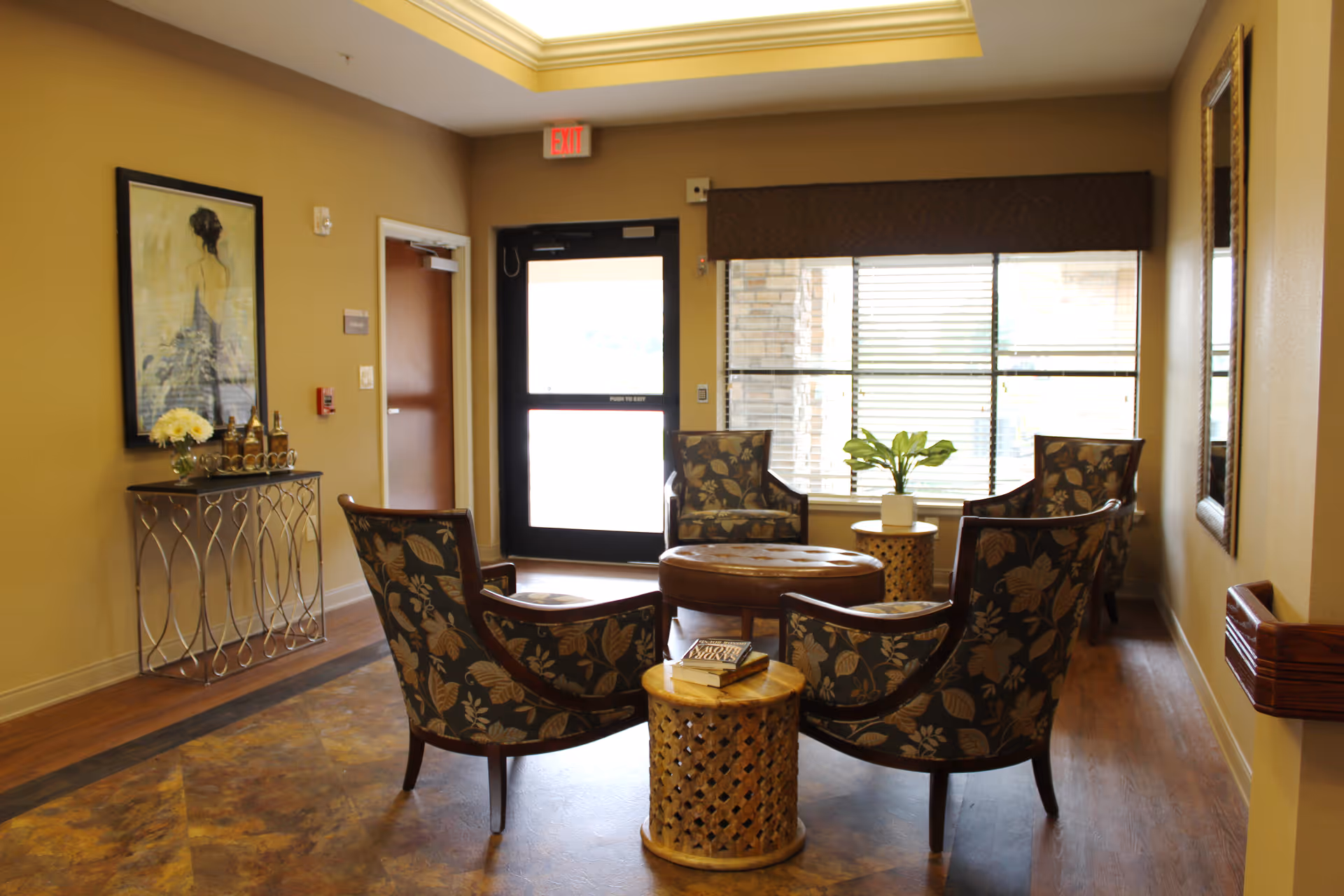 A bright reception lounge with four floral armchairs arranged around a round ottoman and side tables near a window and exit door.