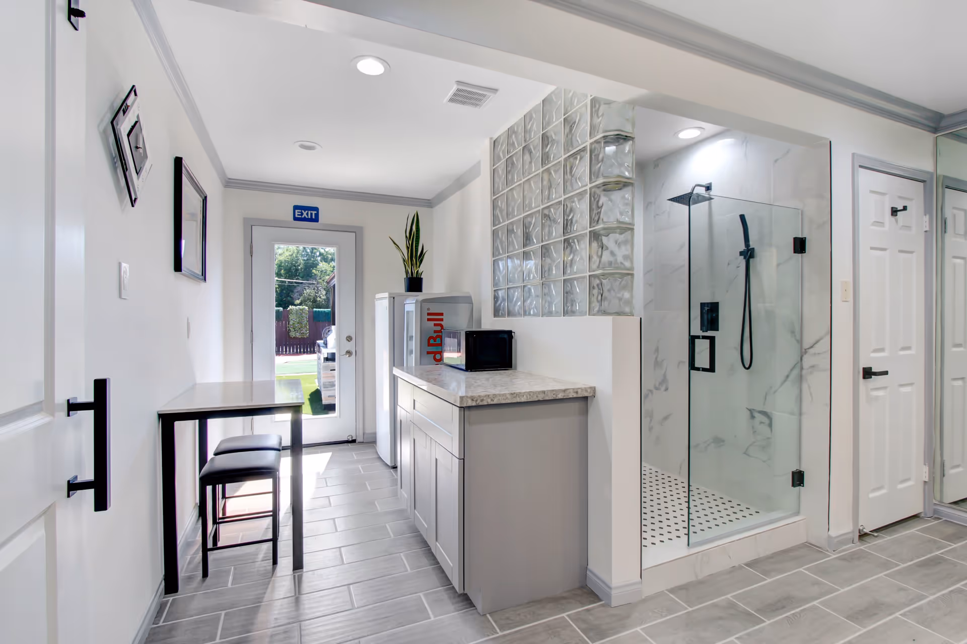 A bright, modern interior space featuring a small kitchenette area with a countertop, microwave, and mini fridge. To the right is a glass-enclosed shower with marble-patterned walls and a black showerhead. A small table with two stools is positioned along the left wall near a door with an exit sign above it, leading outside. The floor is tiled in a light gray color, and the walls are painted white with gray trim.