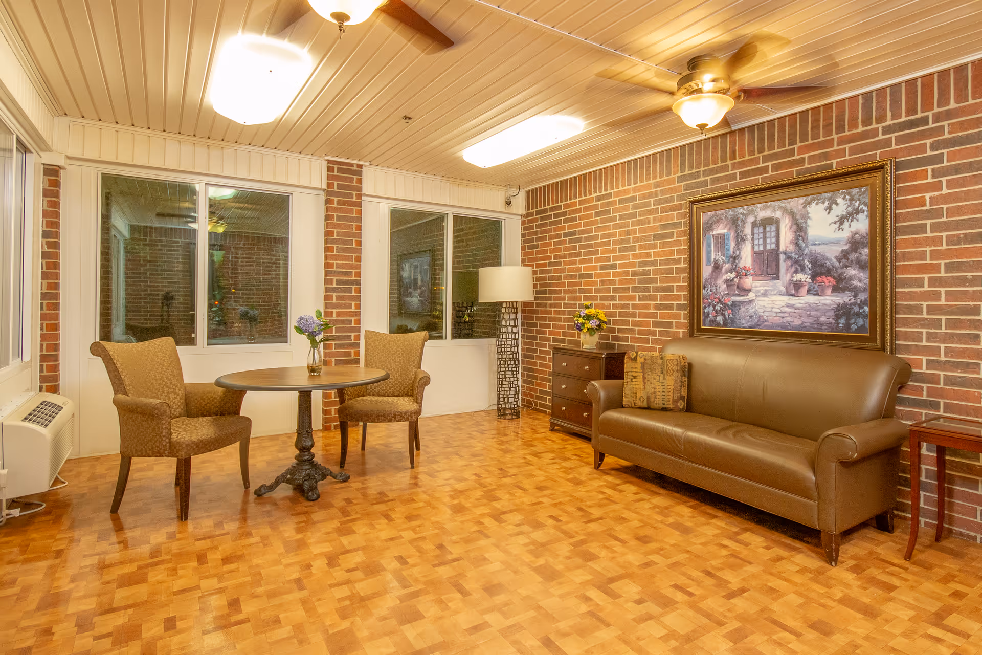 A cozy sitting area with a brown leather sofa, two upholstered armchairs around a small round wooden table with a vase of flowers, a wooden side table, a chest of drawers with a flower vase, a floor lamp, and a large framed painting on a brick wall. The room has parquet flooring, large windows, and ceiling lights with a fan.