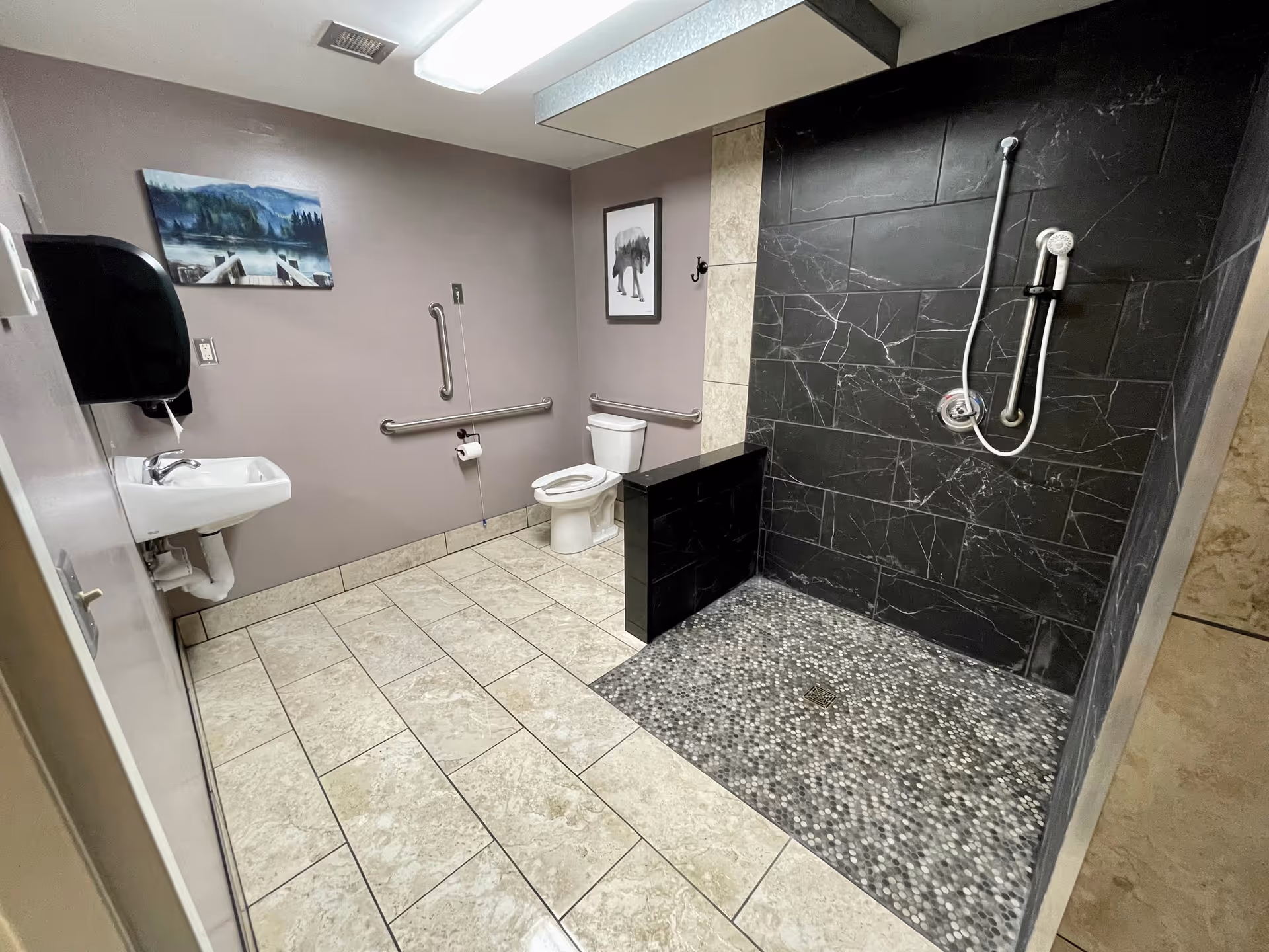 A spacious bathroom with beige tiled floor and walls painted in light brown. The room features a white toilet with grab bars on the wall, a white sink with a black soap dispenser above it, and a walk-in shower area with black marble tiles and a handheld showerhead. Two framed pictures hang on the walls, one depicting a nature scene and the other a bear.