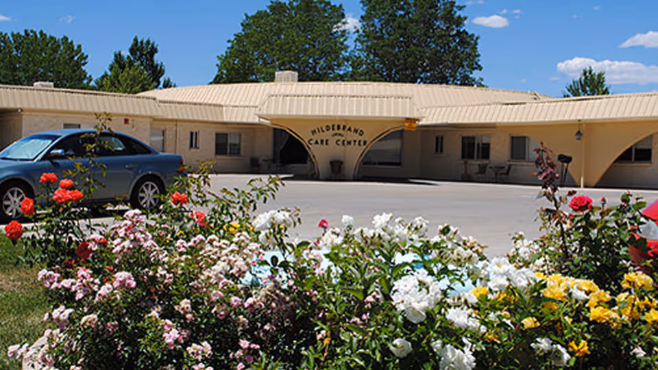 Front entrance of Hildebrand Care Center showing the building facade, colorful flower beds in the foreground, and parked cars.