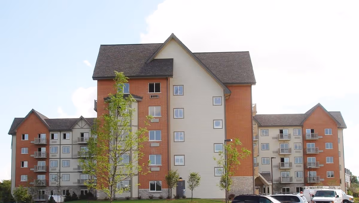 Front exterior of a multi-story residential building with balconies, pitched roofs, and young trees on a lawn.