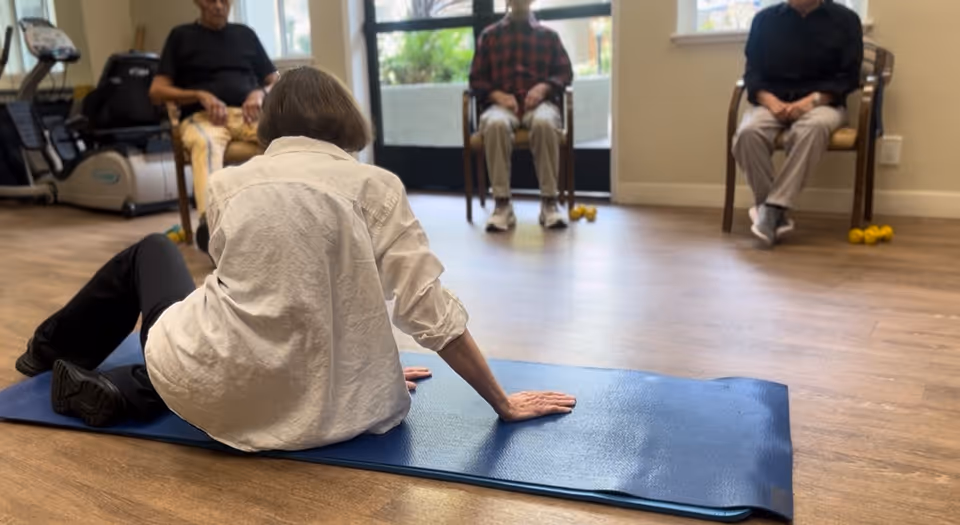 A group of elderly individuals participating in a seated exercise session indoors. One person is on a blue exercise mat on the floor, while three others are seated on chairs in the background. The room has wooden flooring and exercise equipment is visible on the left side.