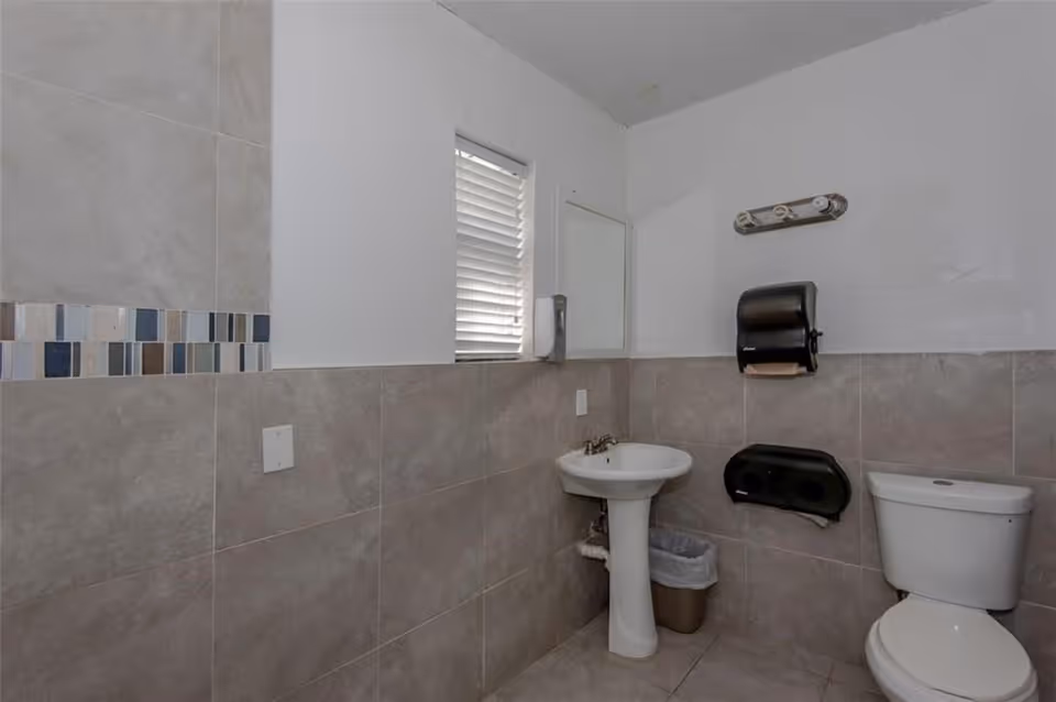 Small restroom with a pedestal sink, toilet, tiled walls, window blinds and wall-mounted paper dispensers.