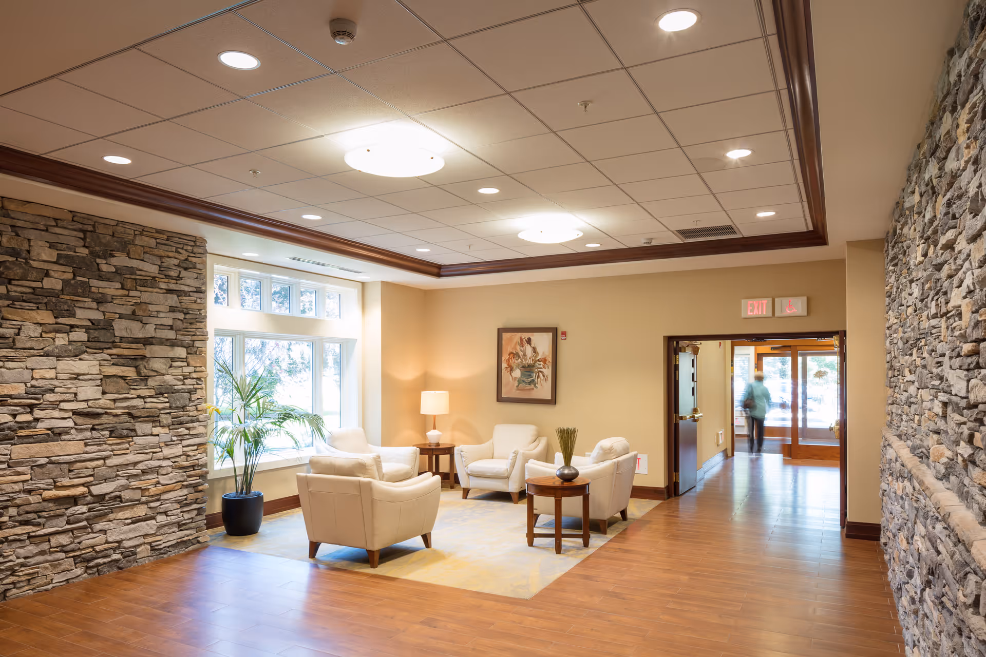 A cozy seating area in a senior living community with four white armchairs arranged around a small wooden table with a vase. The room features stone accent walls, a large window with natural light, a potted plant, a floor lamp, and a framed artwork on the wall. The ceiling has recessed lighting and a decorative wooden trim. In the background, there is a hallway leading to an exit door with a person walking through it.