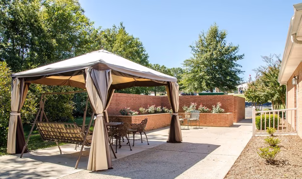 Outdoor patio area with a beige canopy gazebo covering a metal swing and a metal table with chairs. There are brick walls with flower beds and trees in the background under a clear blue sky.