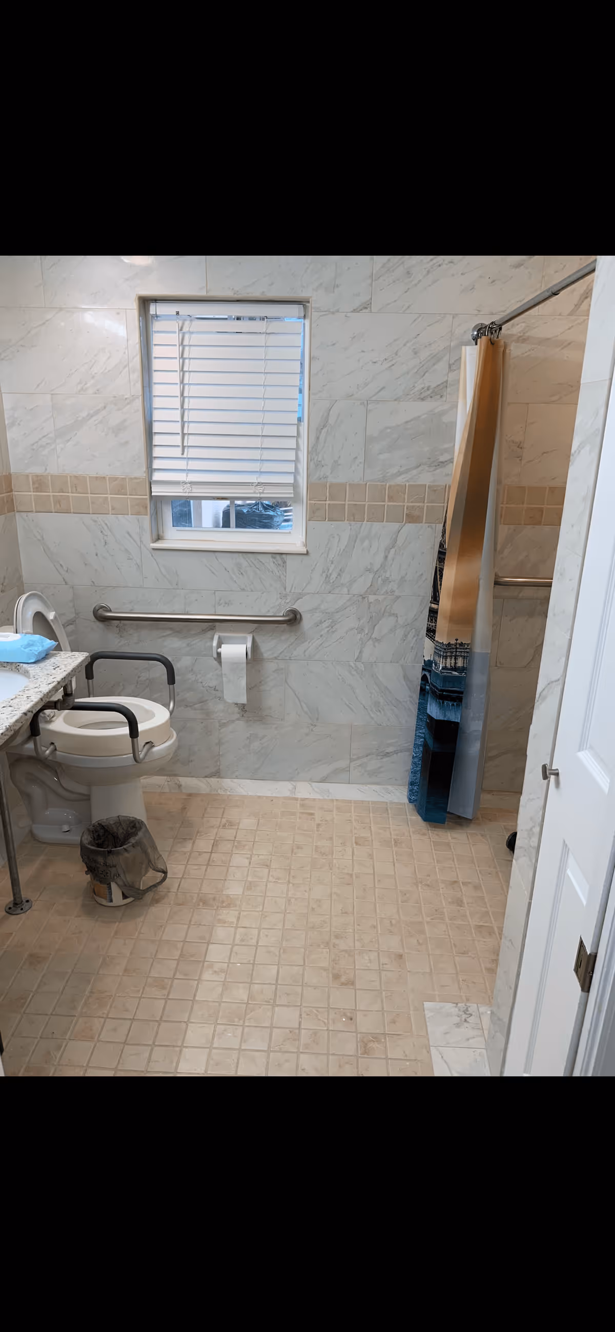 Accessible bathroom with a raised toilet and grab bars, tiled walls and floor, a small window with blinds, and a shower area with a curtain.