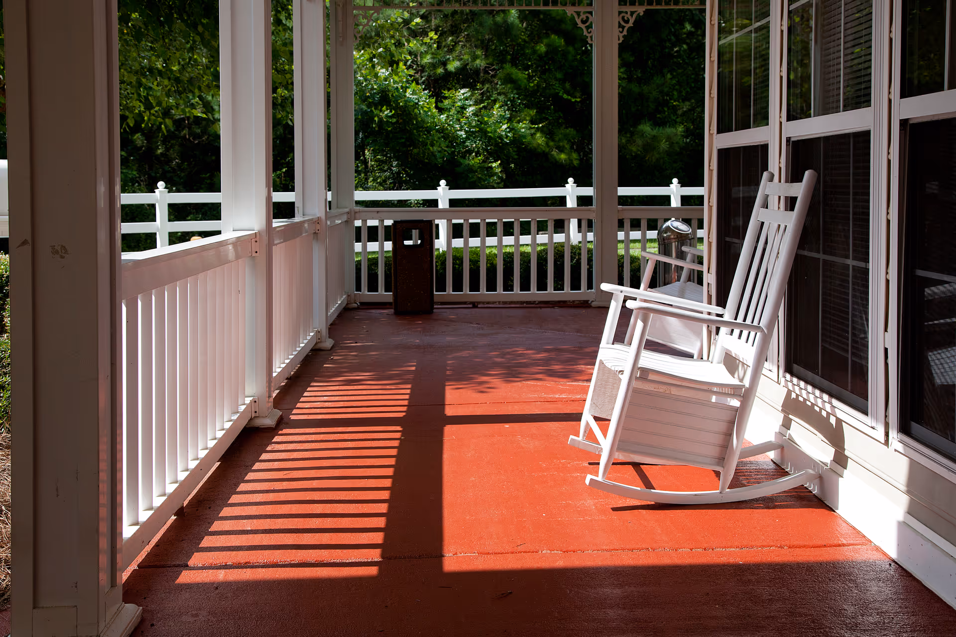A sunlit covered porch with white rocking chairs and a white railing overlooking green trees.
