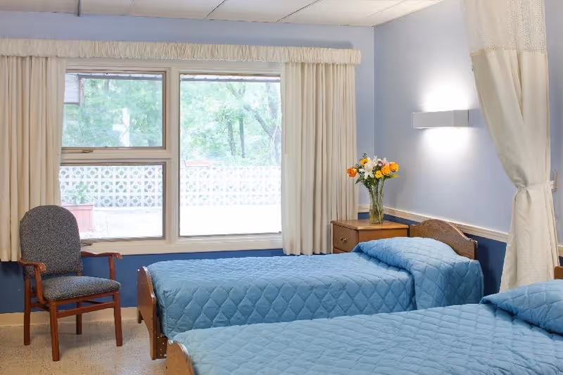 A bright room with two single beds covered in blue quilted bedspreads. Between the beds is a wooden nightstand with a vase of colorful flowers. A wooden chair with a gray cushion is placed near a large window with cream-colored curtains. The walls are painted light blue and a wall-mounted light fixture is above the nightstand.