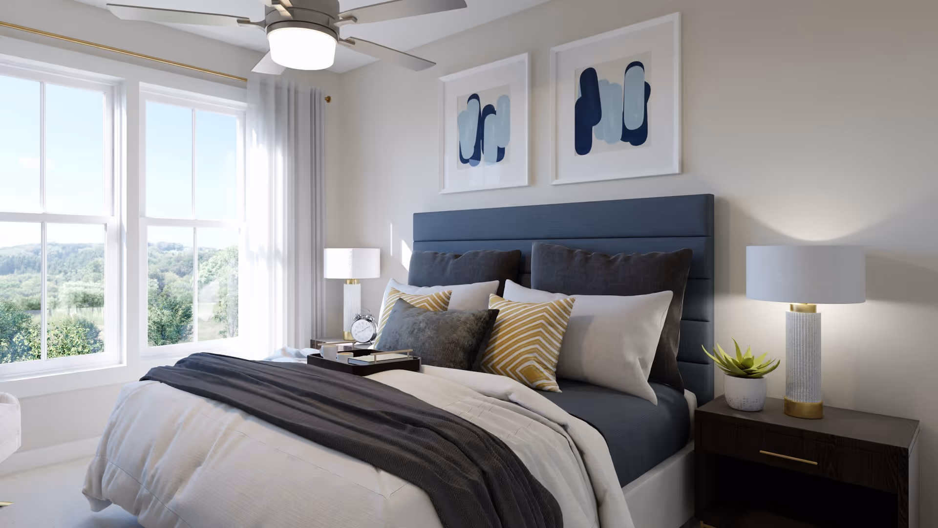 A bright bedroom with a large window showing a green outdoor view. The bed has a blue upholstered headboard and is made with white and dark gray bedding, along with multiple pillows in shades of gray and yellow. There are two framed abstract artworks above the bed. On each side of the bed, there are dark wooden nightstands with modern lamps and a small plant on the right nightstand. A ceiling fan with a light is mounted on the ceiling.