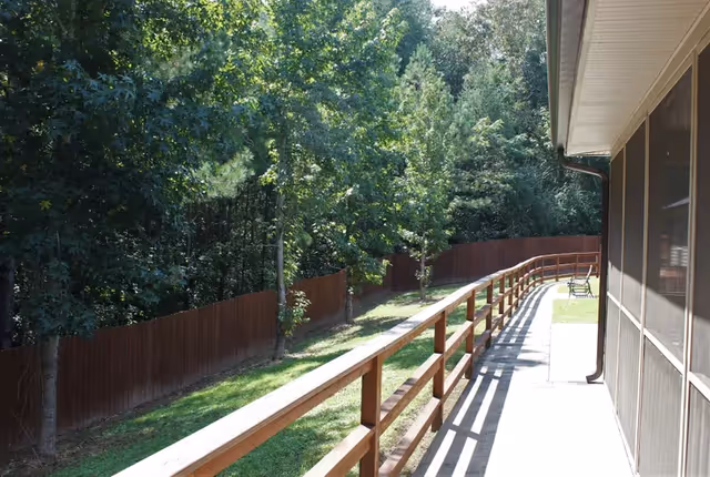 A sunny outdoor walkway alongside a building with a wooden railing and a brown fence. Trees and greenery are visible beyond the fence, and a bench is placed near the end of the walkway.