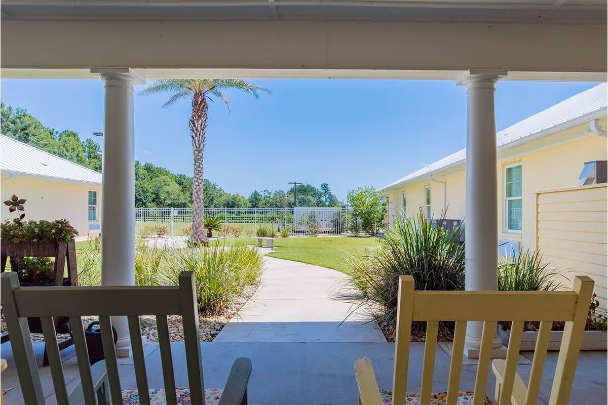 View from a covered porch with rocking chairs looking out onto a landscaped courtyard with a palm tree, bushes, a paved walkway, and two single-story buildings under a clear blue sky.