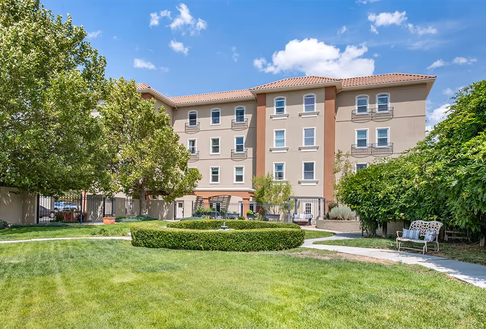 Exterior view of a multi-story senior living facility building with beige walls and red-tiled roof, surrounded by green trees, bushes, and a well-maintained lawn with a circular hedge and a small fountain in the center. There is a paved walkway and a bench with cushions on the right side under the trees.