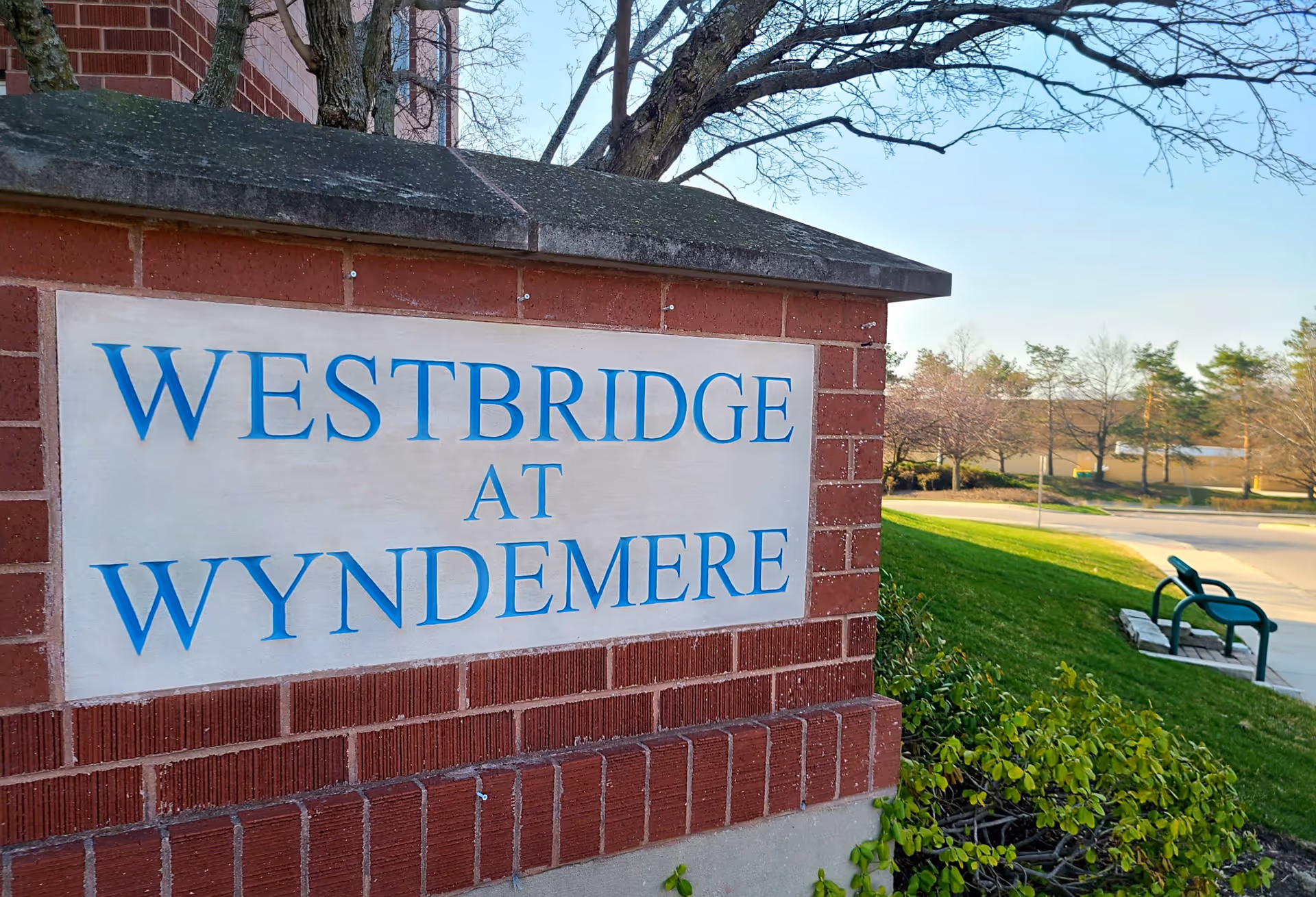 A brick sign with blue letters reading 'WESTBRIDGE AT WYNDEMERE' situated outdoors with a grassy area, trees, and a bench visible in the background under a clear sky.