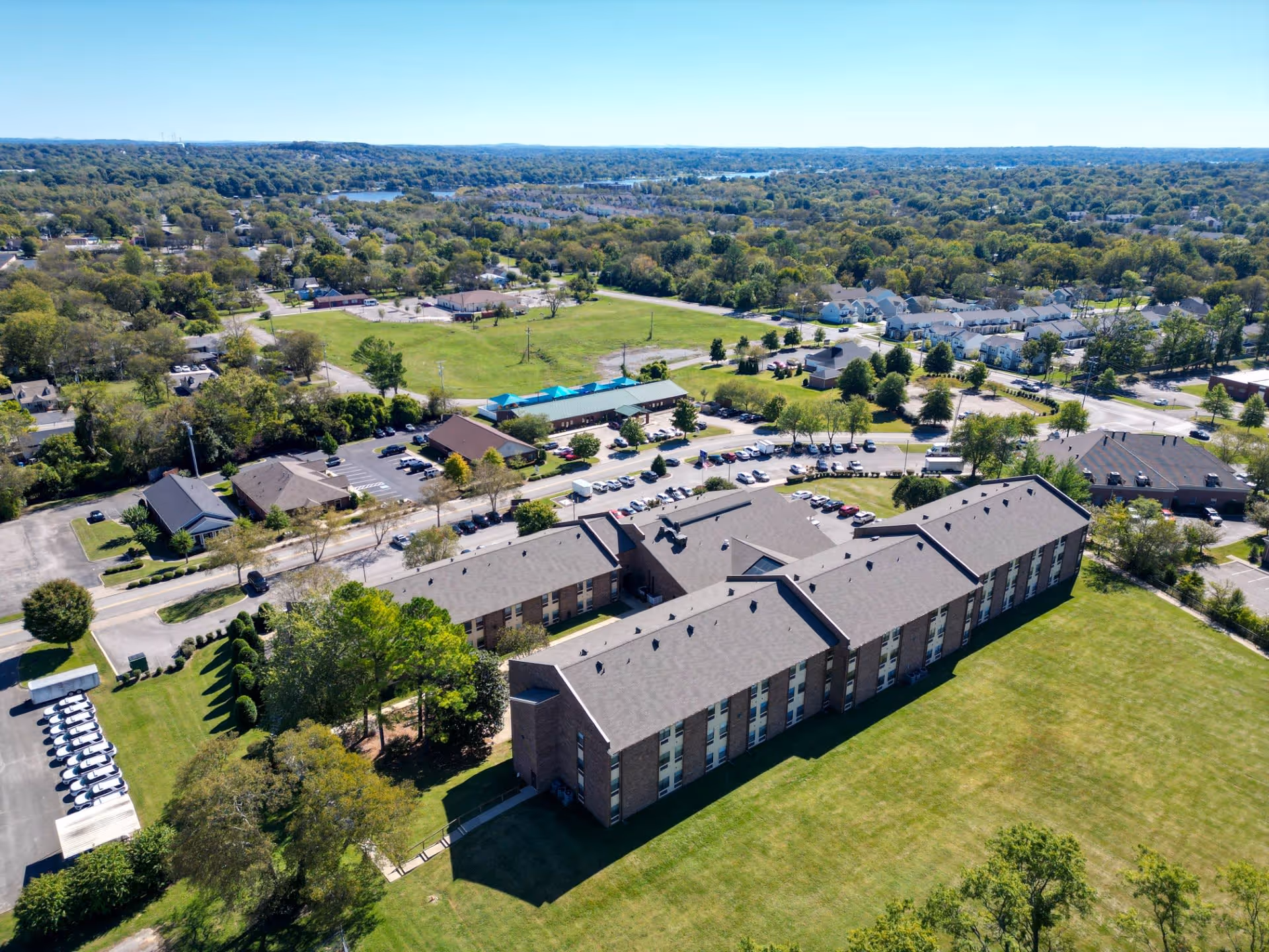 Aerial view of Park Place Retirement Community showing a large multi-wing building surrounded by green lawns, trees, parking lots, and nearby residential areas under a clear blue sky.