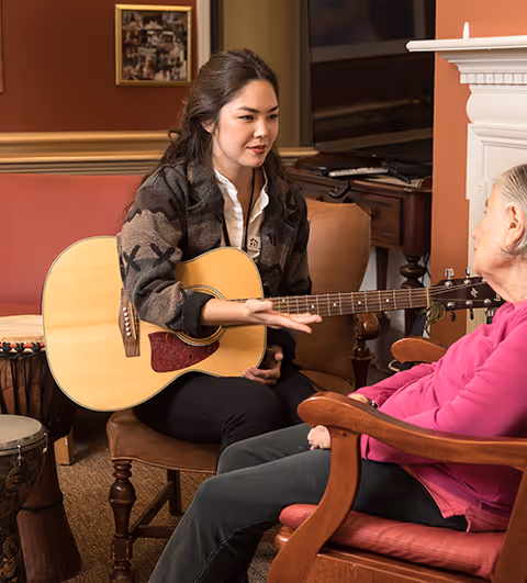 A young woman sitting on a chair playing an acoustic guitar and engaging with an elderly woman seated across from her in a cozy living room setting with warm lighting and comfortable furniture.