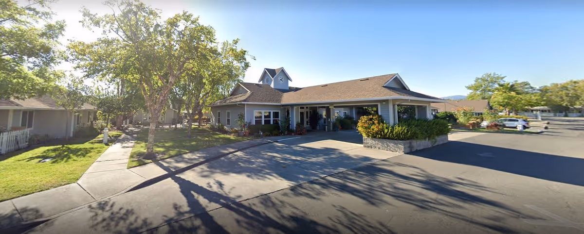 Exterior view of Sonoma Hills Retirement Living Community showing a single-story building with a covered entrance, surrounded by trees, greenery, and a paved driveway under a clear blue sky.