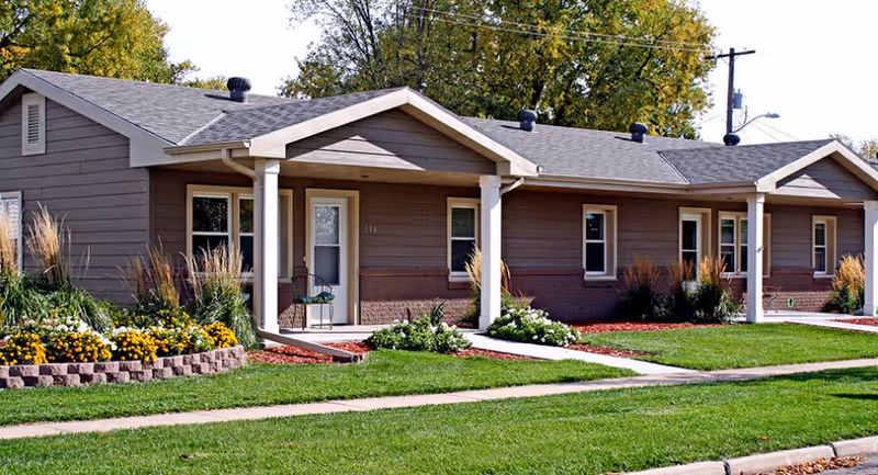 Single-story residential building with brown siding and white trim, featuring multiple entrances with small porches. The front yard is landscaped with green grass, flower beds with yellow and white flowers, and ornamental grasses. Trees with green foliage are visible in the background under a clear sky.