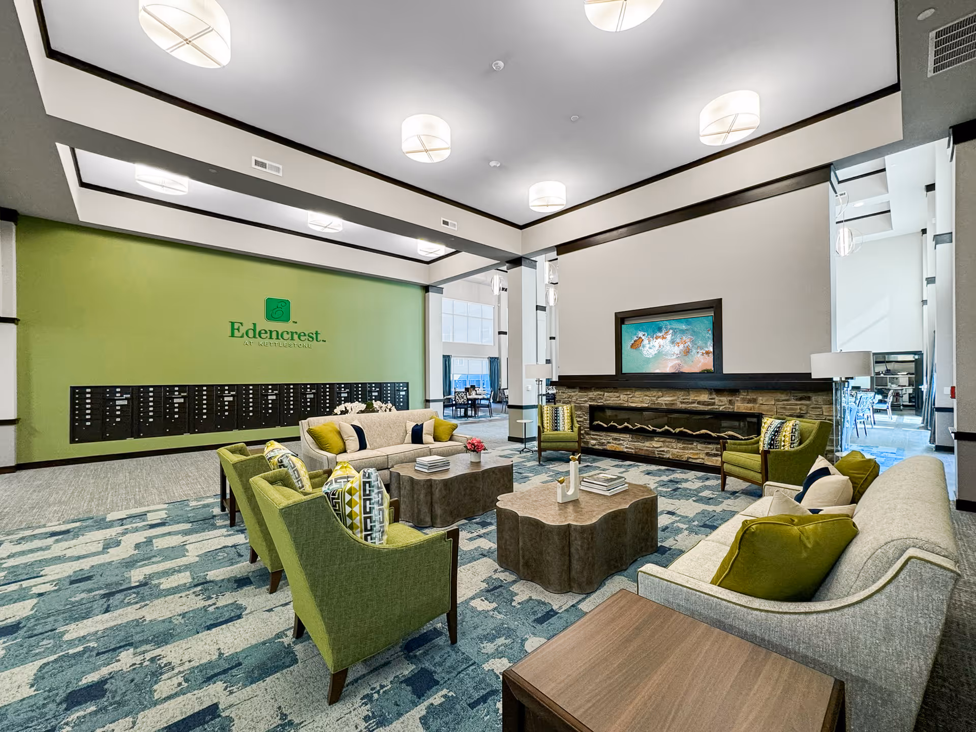 A spacious and modern living room area in Edencrest at Kettlestone featuring comfortable green and beige sofas and armchairs arranged around two uniquely shaped coffee tables. The room has a patterned blue and beige carpet, a stone fireplace with a colorful abstract painting above it, and a green accent wall with the Edencrest at Kettlestone logo and mailboxes. Large windows and contemporary ceiling lights brighten the space.
