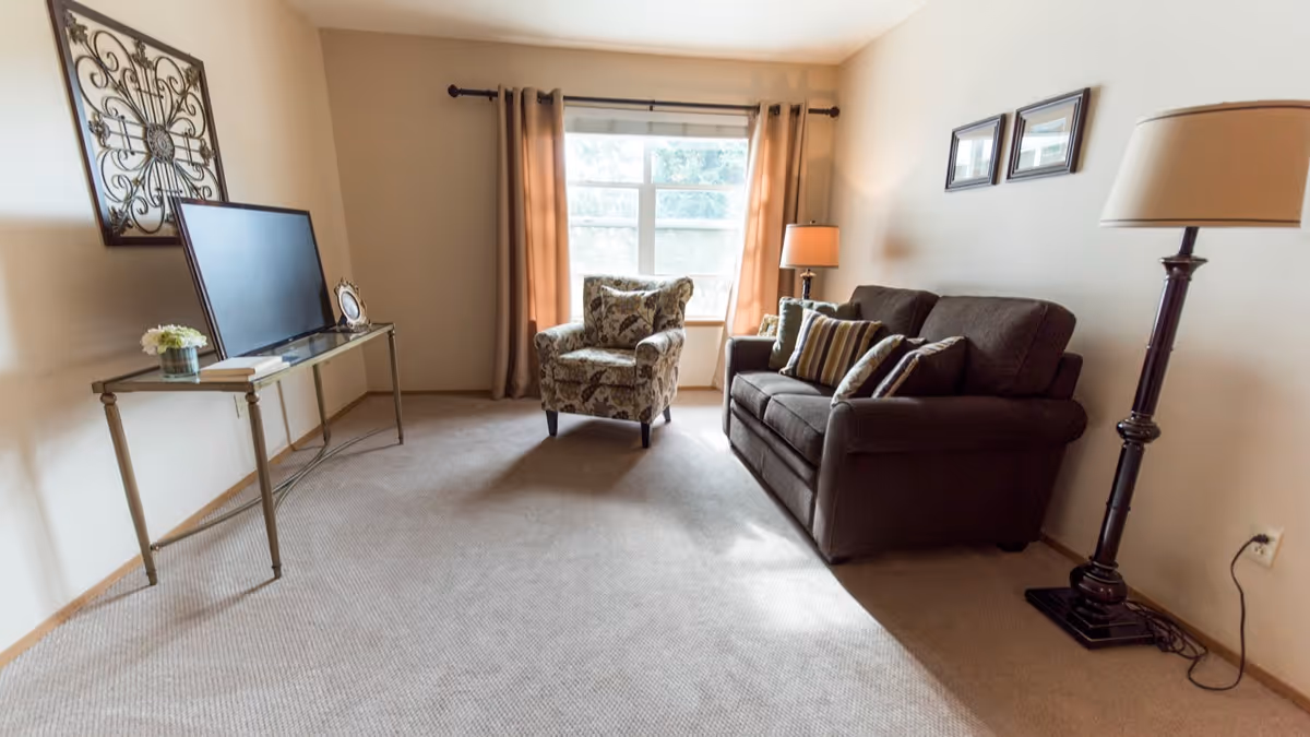 A cozy living room with a dark brown sofa adorned with striped pillows, a patterned armchair near a window with beige curtains, a floor lamp, and a glass console table holding a flat-screen TV, a small plant, and a picture frame. The room has beige walls and carpeted flooring.