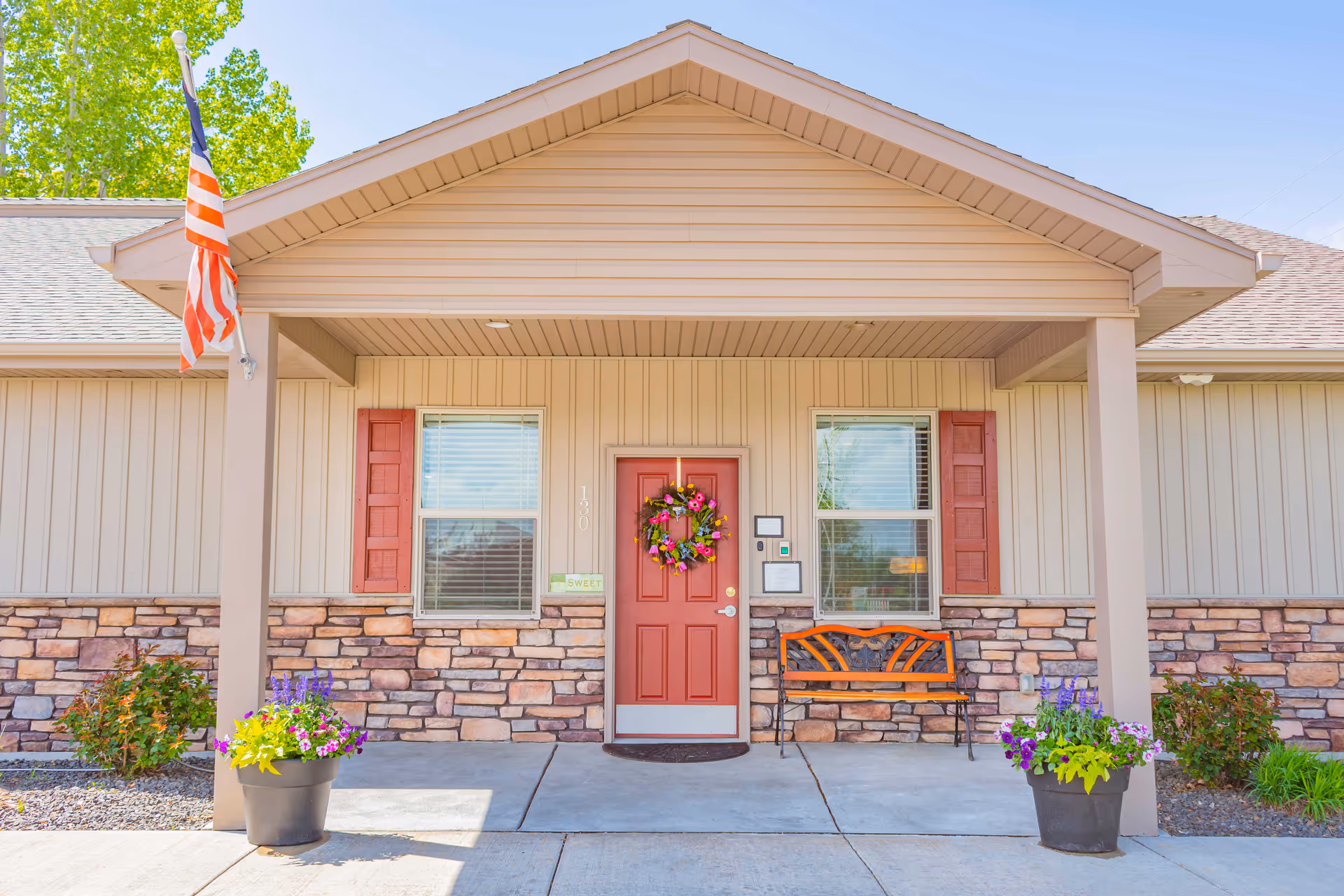 Front entrance of a single-story building with a red door adorned with a wreath, a bench, potted flowers, and an American flag.