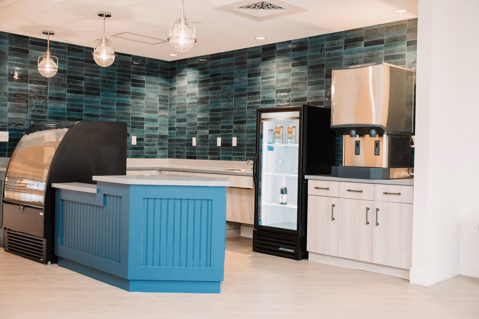 A modern kitchen area with a blue island counter, a refrigerated display case with beverages, a stainless steel water dispenser, light wood cabinets, and dark green tiled walls under hanging glass pendant lights.